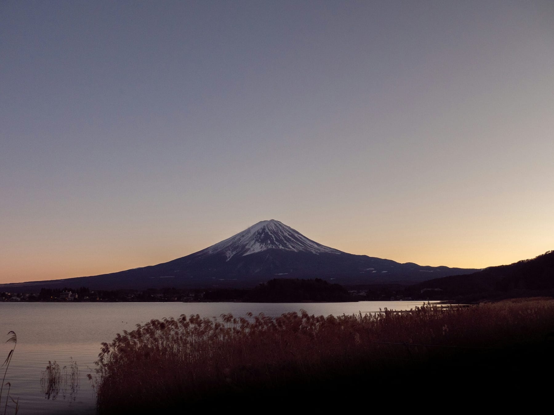 Snow-capped Mount Fuji reflected in a still lake at dusk, warm orange sky fading to purple above the perfect cone, reed grasses in the foreground