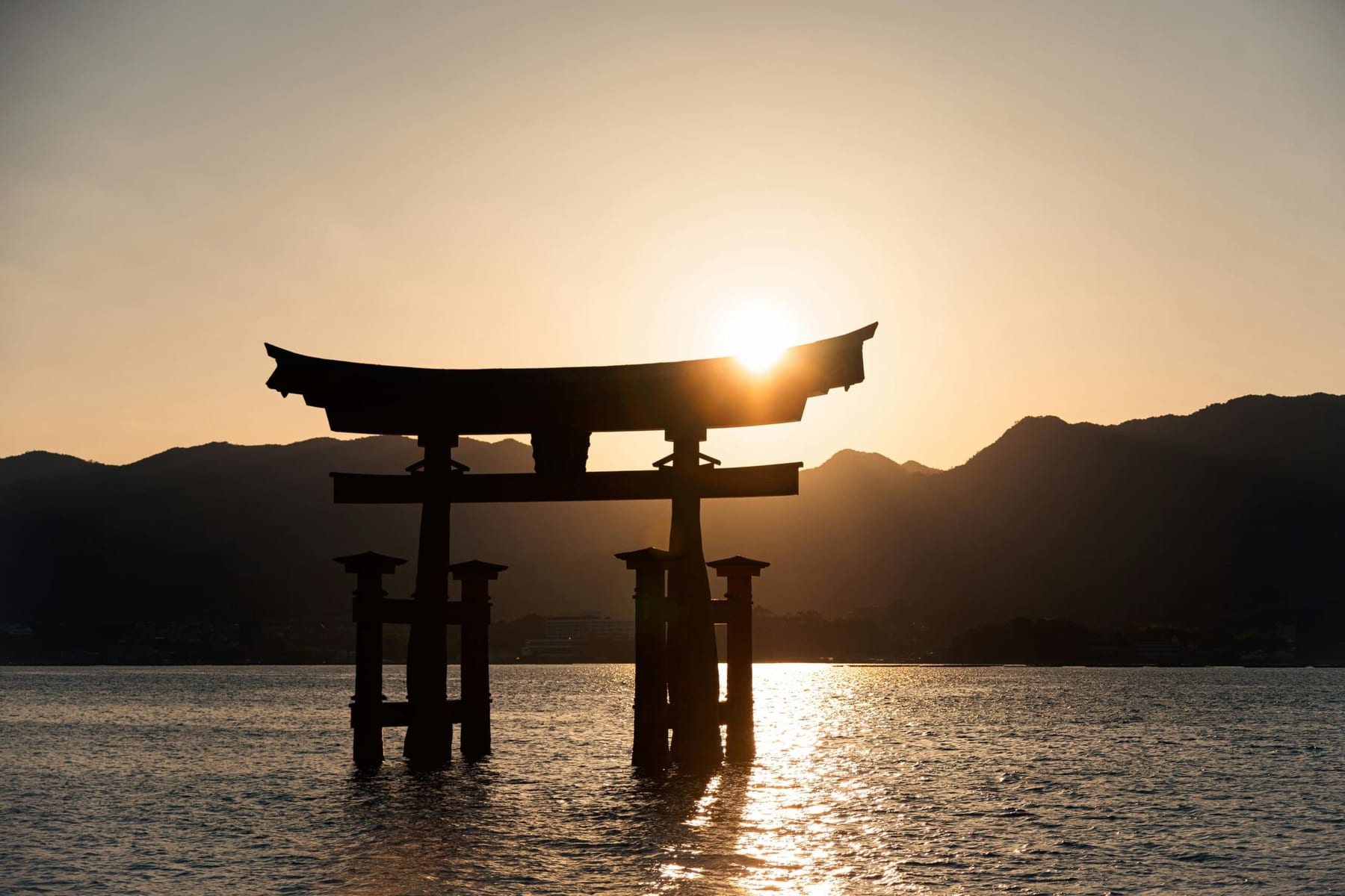 The torii gate of Hakone Shrine silhouetted in gold against a sunset sky, standing in the water with mountains behind