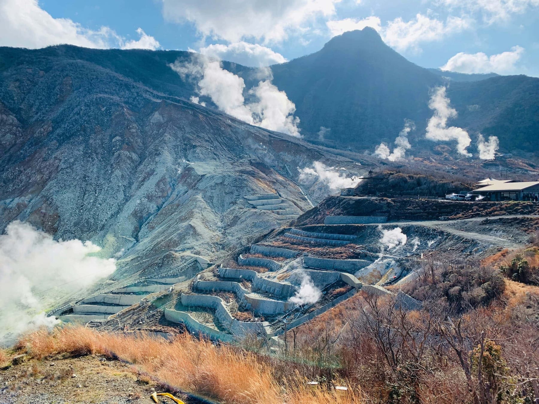 Owakudani volcanic valley in Hakone, sulphurous steam rising from multiple vents across the rock terraces, the volcanic mountain rising behind under a blue sky