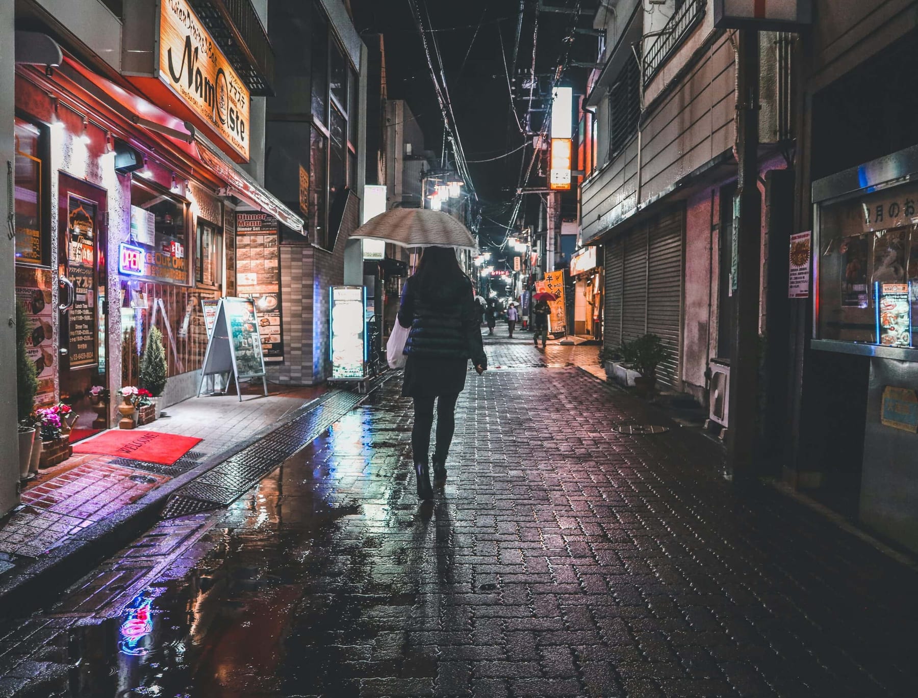 A woman in a dark jacket walks alone down a rain-soaked Japanese alley at night, holding a white umbrella, neon lights reflected in the pavement