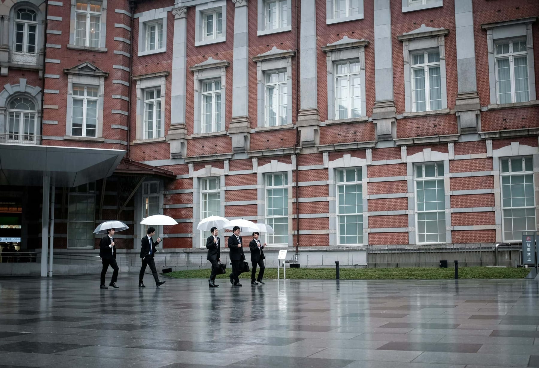 Businessmen in dark suits carrying white umbrellas cross the plaza in front of Tokyo Station's red-brick Marunouchi facade on a grey rainy day