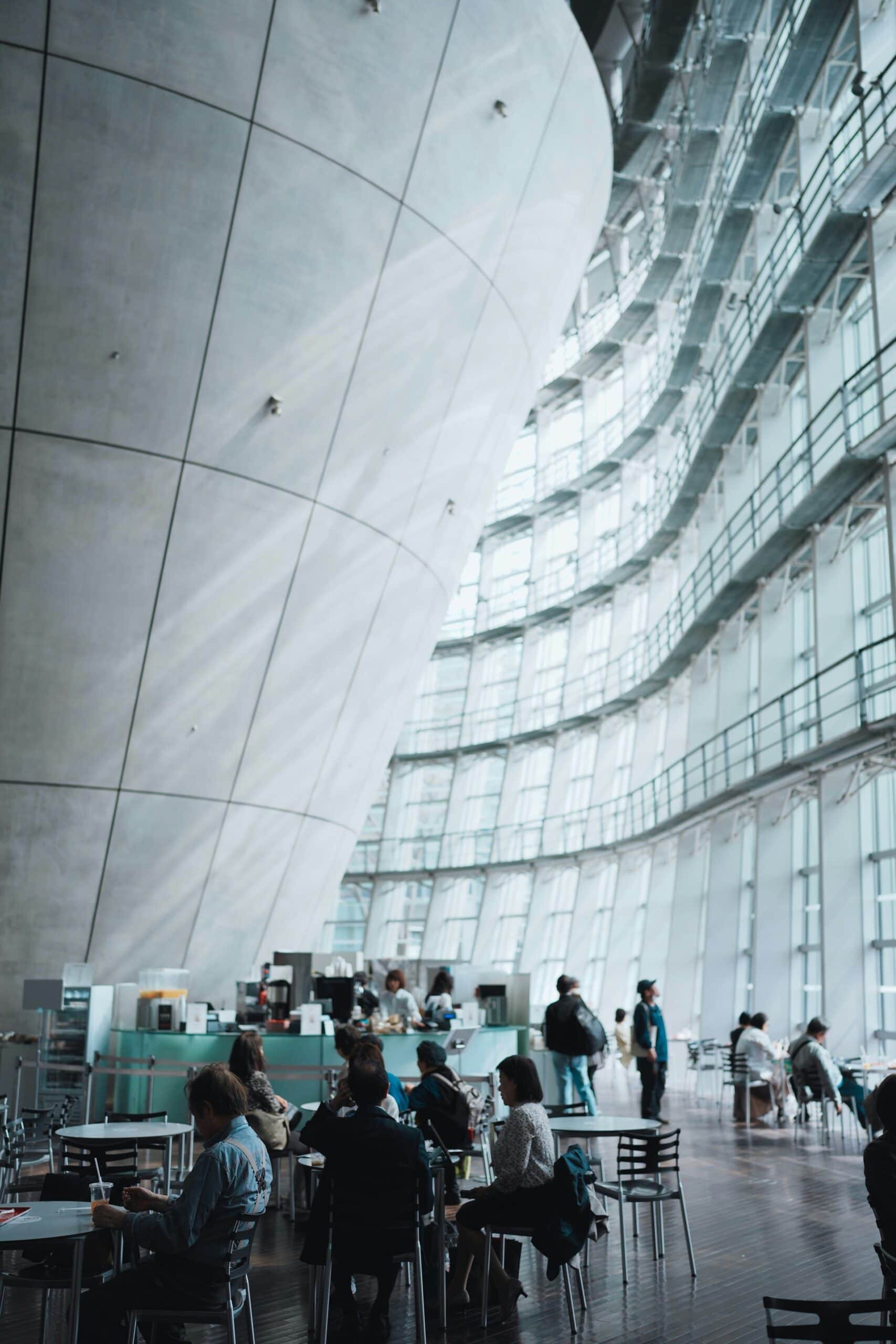 Visitors seated inside the vast curved glass atrium of a contemporary Japanese museum, calm and luminous on a rainy day trip from Tokyo